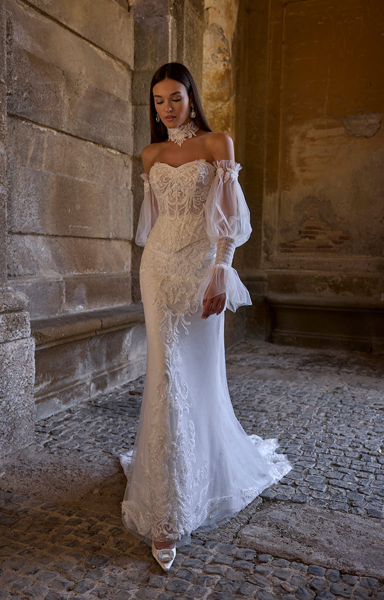 Woman in 63207 Atlas by Veni Infantino Bridal - Off-the-Shoulder Lace Wedding Dress with Sheer Puff Sleeves standing against a stone wall.