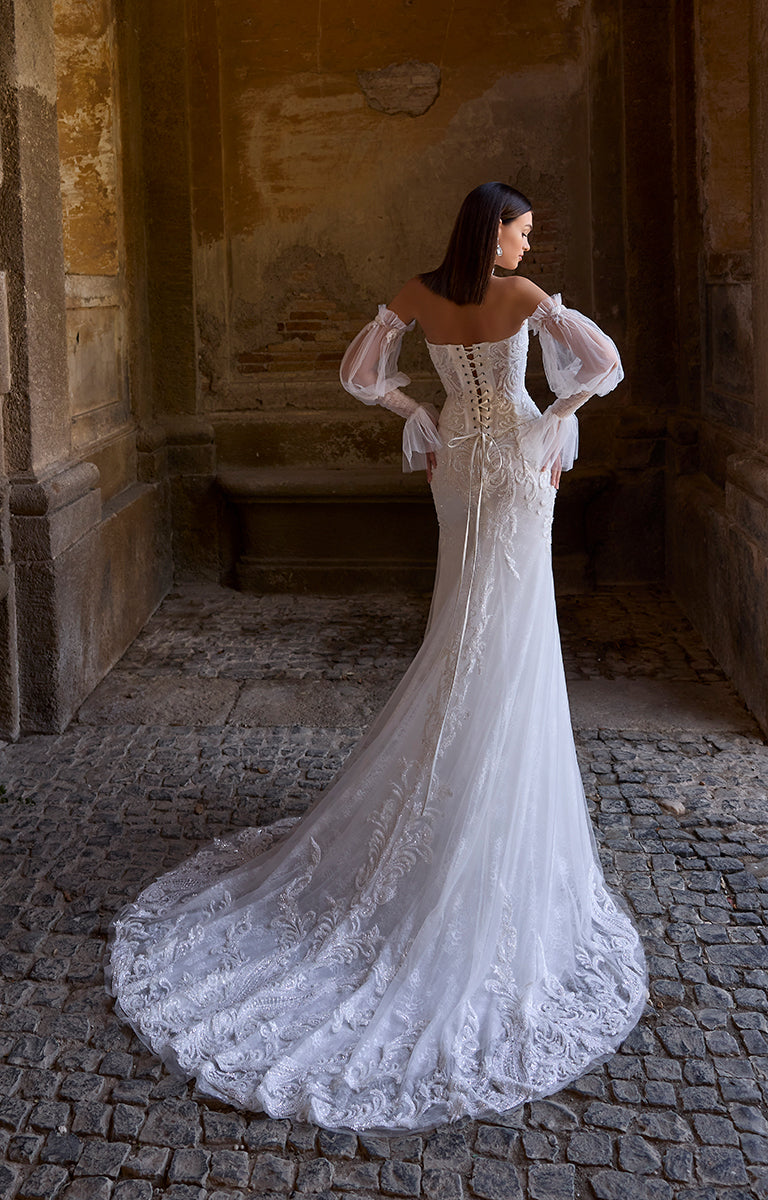 Woman in 63207 Atlas by Veni Infantino Bridal - Off-the-Shoulder Lace Wedding Dress with Sheer Puff Sleeves standing in a stone archway.