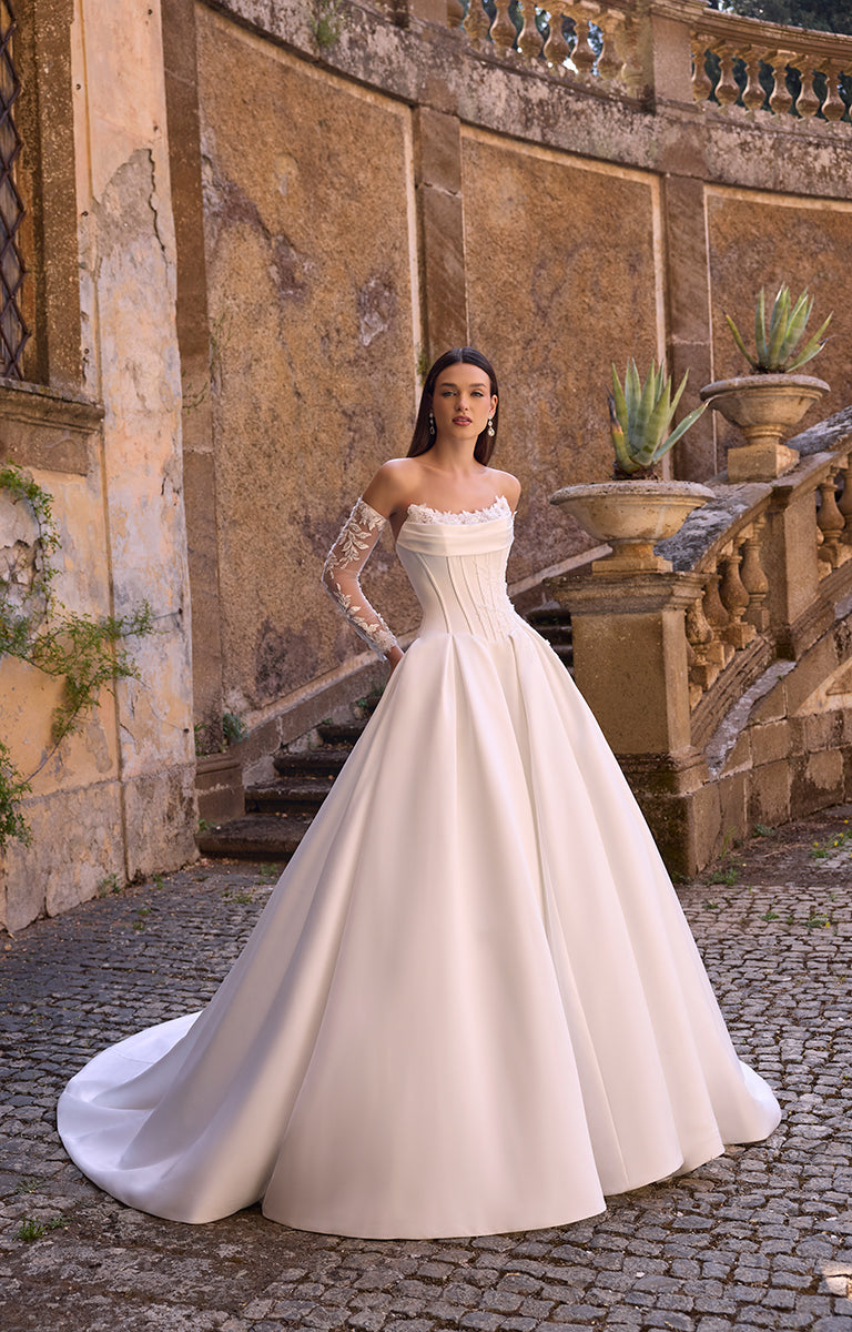 Woman in 63228 Annie by Veni Infantino Bridal - Strapless Corset Ball Gown with Detachable Lace Sleeves standing in front of an ornate stone wall.