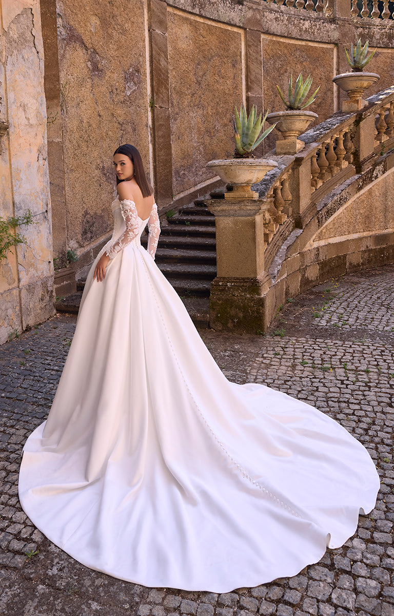 Woman in 63228 Annie by Veni Infantino Bridal - Strapless Corset Ball Gown with Detachable Lace Sleeves standing in an elegant outdoor setting with stone walls and steps.