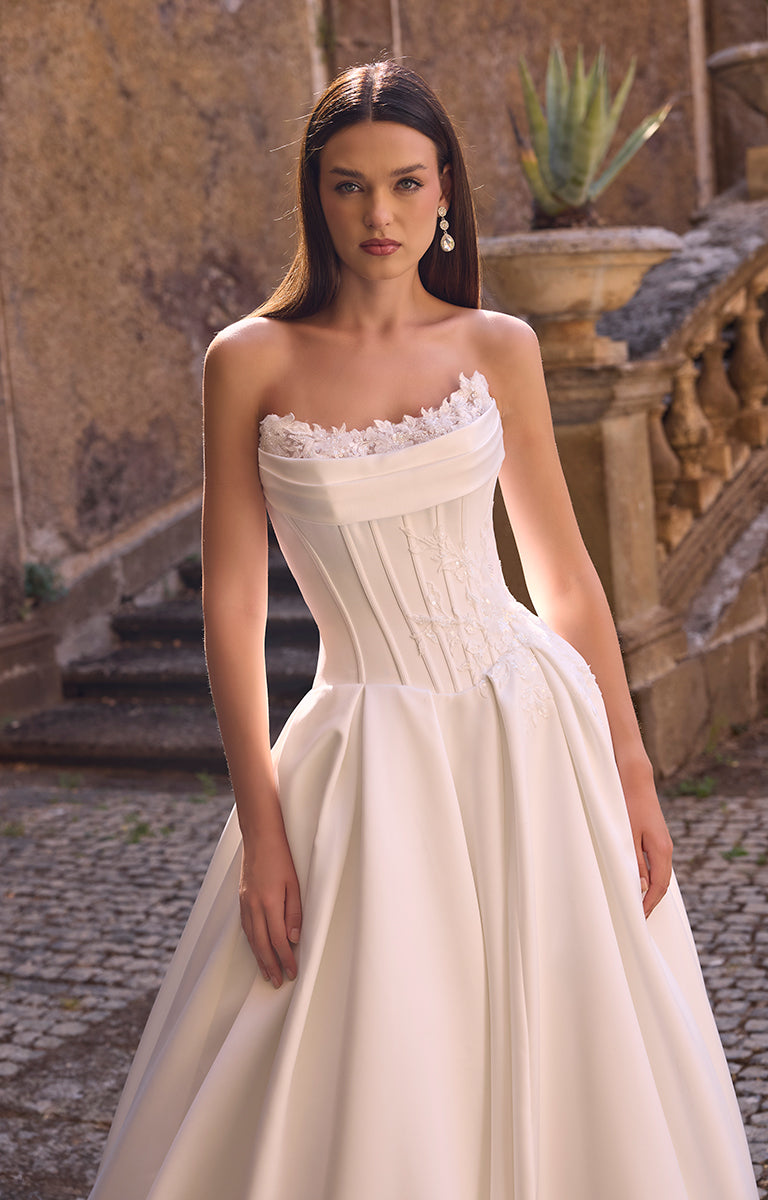 Woman in 63228 Annie by Veni Infantino Bridal - Strapless Corset Ball Gown with Detachable Lace Sleeves removed standing outdoors with classical architecture in the background.