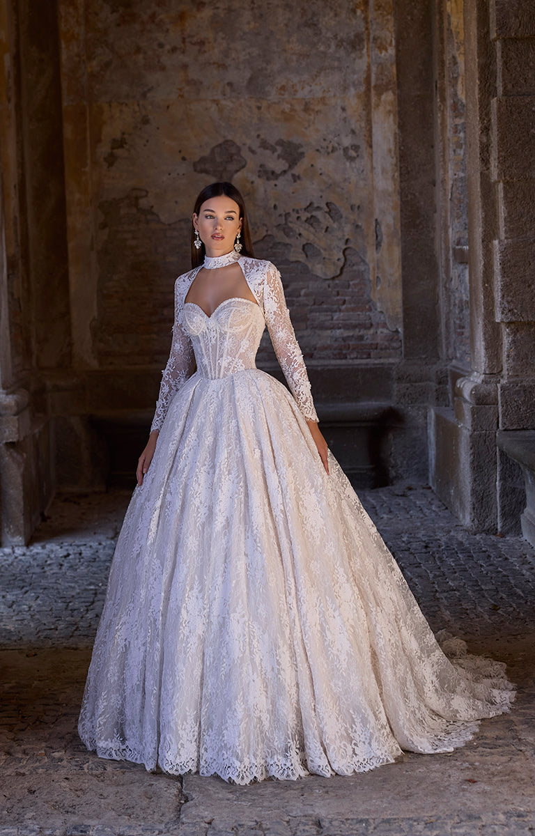 Woman in 63241 Aavya by Veni Infantino Bridal - Lace Ballgown with High Neckline and Illusion Sleeves standing in a stone architectural setting