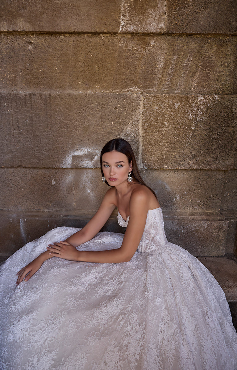 Woman in 63241 Aavya by Veni Infantino Bridal - Lace Ballgown with High Neckline and Illusion Sleeves sitting against a stone wall.