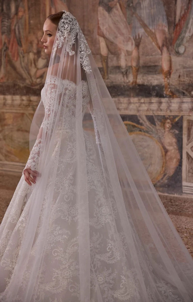 A woman wearing Woman wearing Veni Infantino 63325 Alagna - Lace Ball Gown Wedding Dress with Off-the-Shoulder Neckline, Long Sleeves and Cathedral Train against a the background of an ornately decorated mural in the room of a fine stately home and a matching long lace-trimmed veil, posed in front of a classical fresco wall.