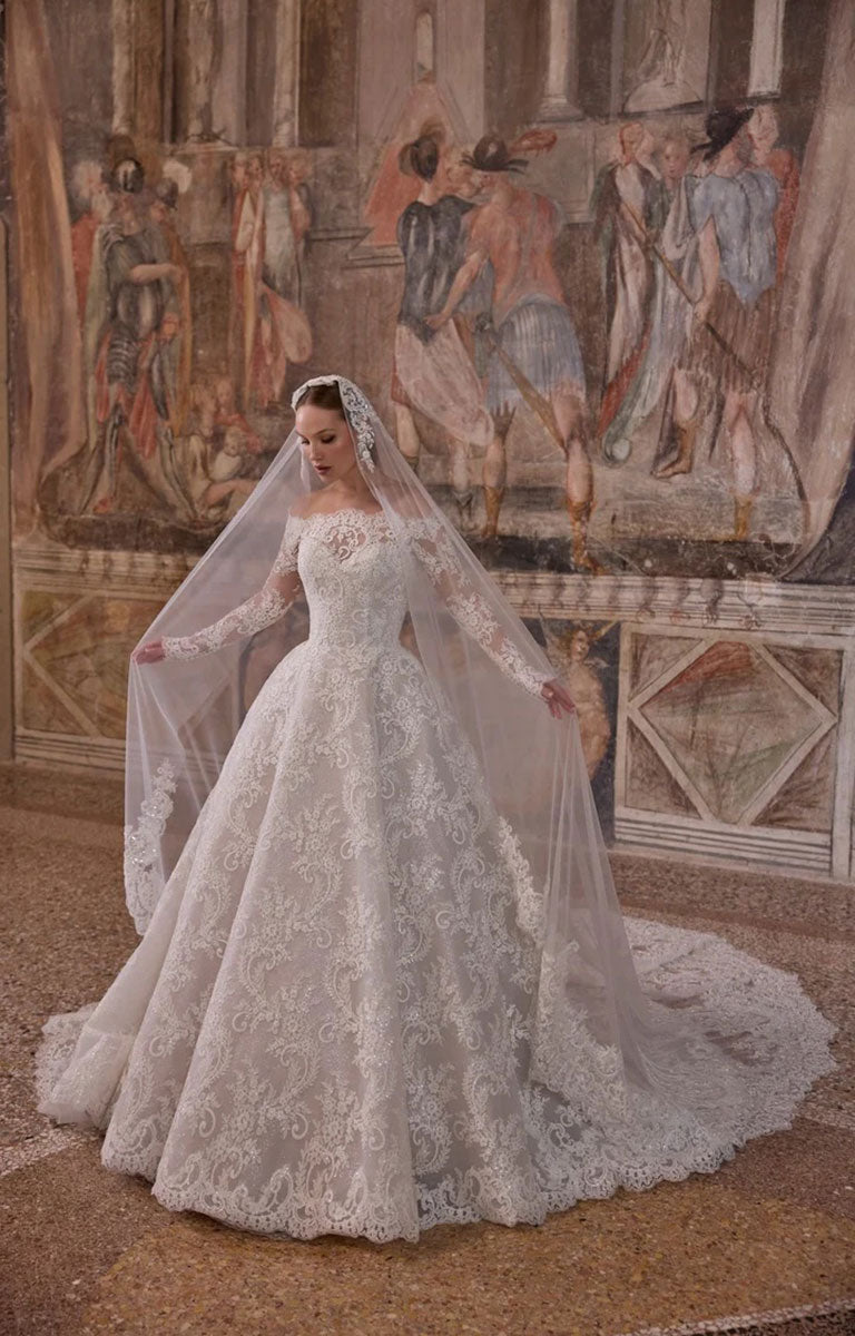 A woman wearing Woman wearing Veni Infantino 63325 Alagna - Lace Ball Gown Wedding Dress with Off-the-Shoulder Neckline, Long Sleeves and Cathedral Train against a the background of an ornately decorated mural in the room of a fine stately home and a long veil, standing in a grand interior with a painted mural background.