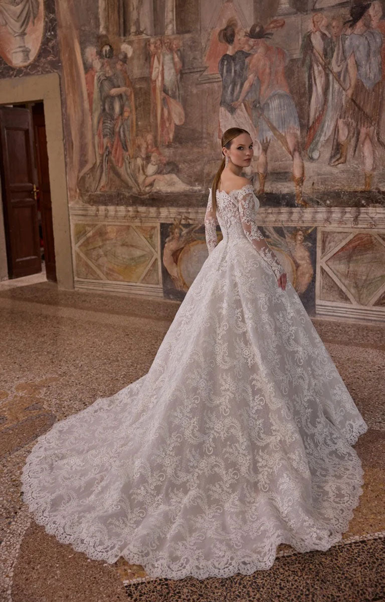 Woman wearing Veni Infantino 63325 Alagna - Lace Ball Gown Wedding Dress with Off-the-Shoulder Neckline, Long Sleeves and Cathedral Train against a the background of an ornately decorated mural in the room of a fine stately home. 