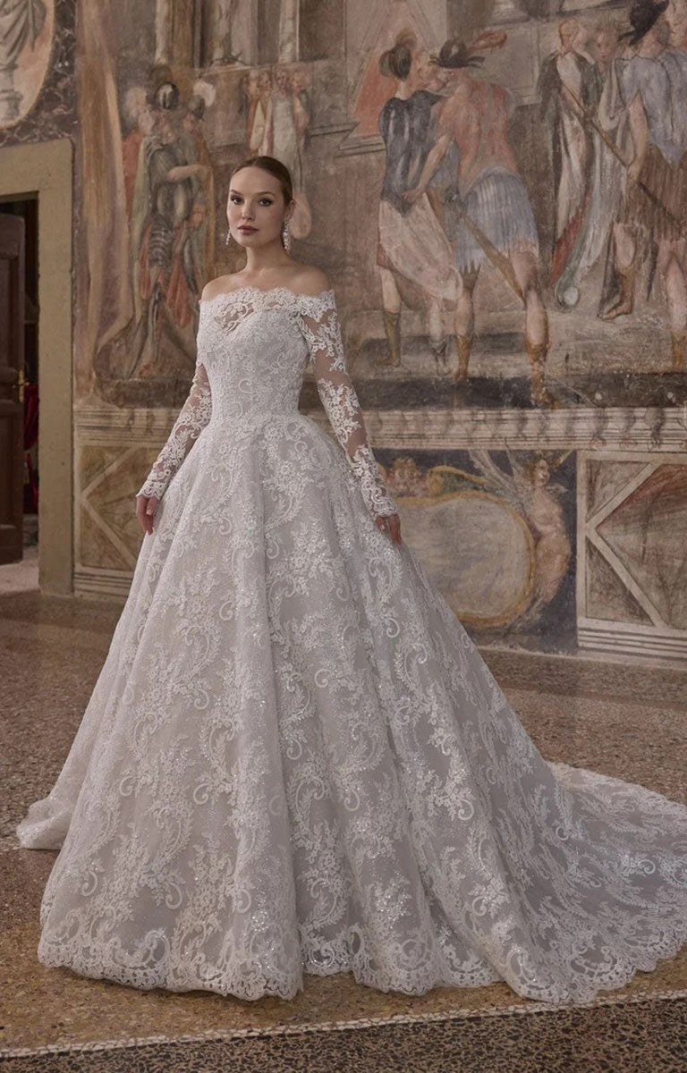 Woman wearing Veni Infantino 63325 Alagna - Lace Ball Gown Wedding Dress with Off-the-Shoulder Neckline, Long Sleeves and Cathedral Train against a the background of an ornately decorated mural in the room of a fine stately home. 