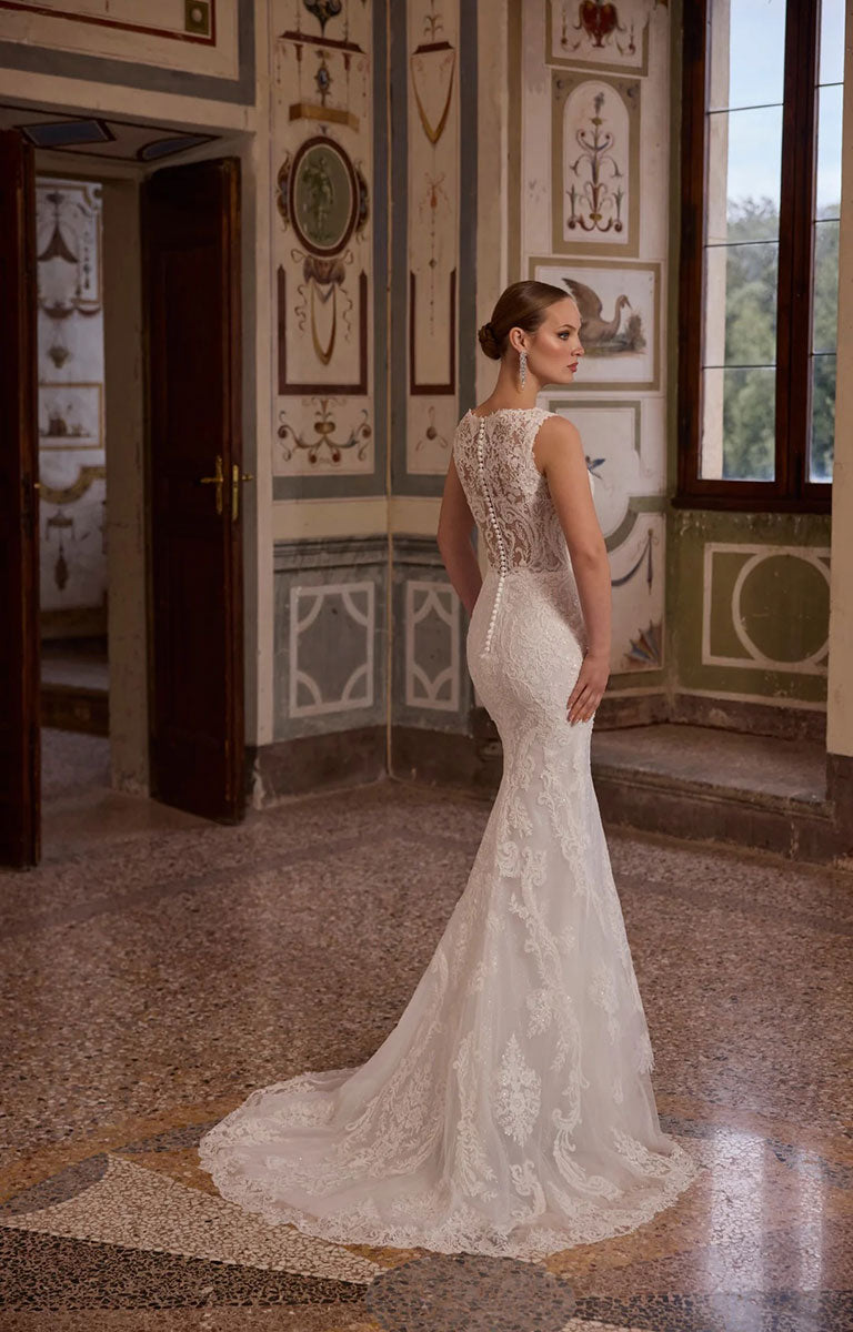 A woman wearing Veni Infantino Algarve 63332 - Fitted Lace Wedding Dress with High Illusion Neckline, Sparkle and Detachable Belt stands in a grand interior room with patterned walls and a large window, shown from the back with the train of the dress visible on the floor.