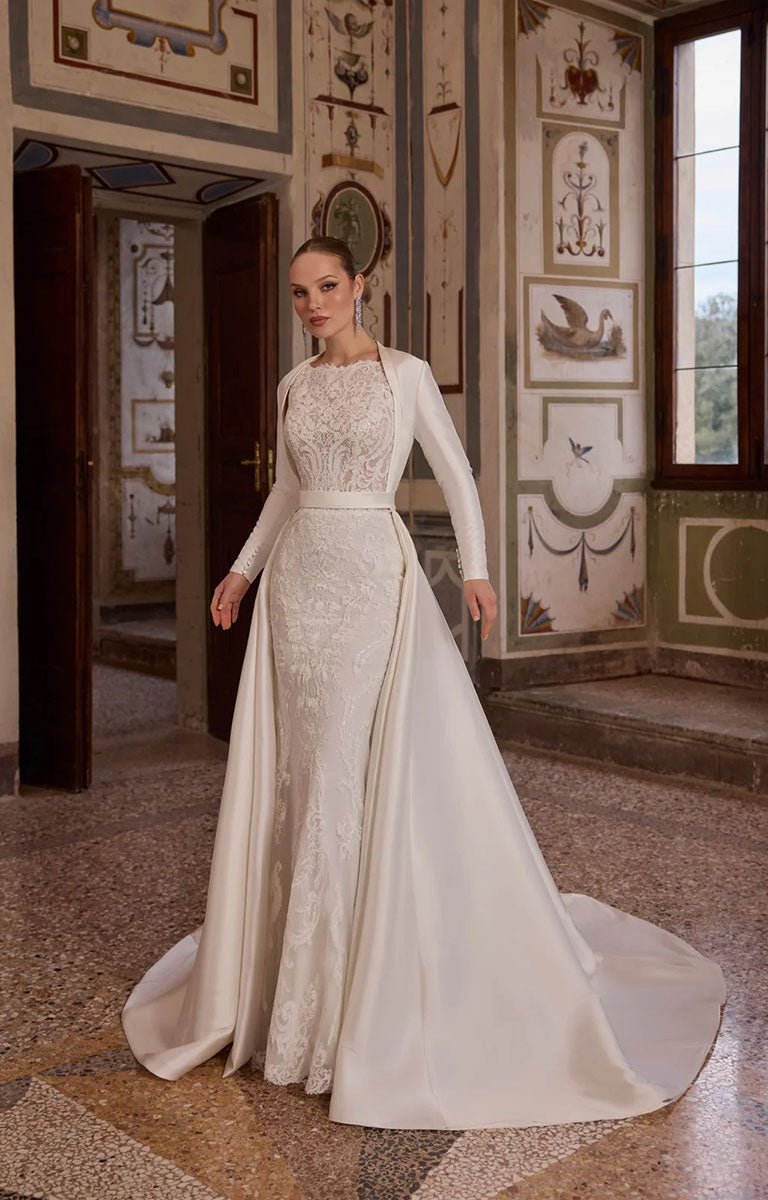 A woman modelling Veni Infantino Algarve 63332 - Fitted Lace Wedding Dress with High Illusion Neckline, Sparkle and Detachable Belt featuring optional Coat 10370 standing in an ornate historic room with decorative walls and windows.