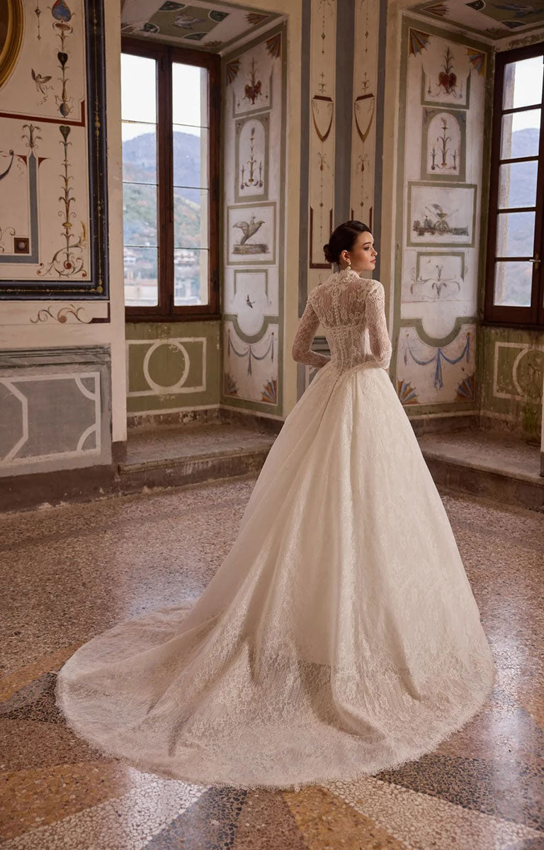 A woman wearing a Alyx by Veni Infantino Bridal - All-Over Lace Ball Gown with High Neckline and Illusion Sleeves with a full ball gown skirt and train, standing in an ornate interior room with decorative walls and windows.