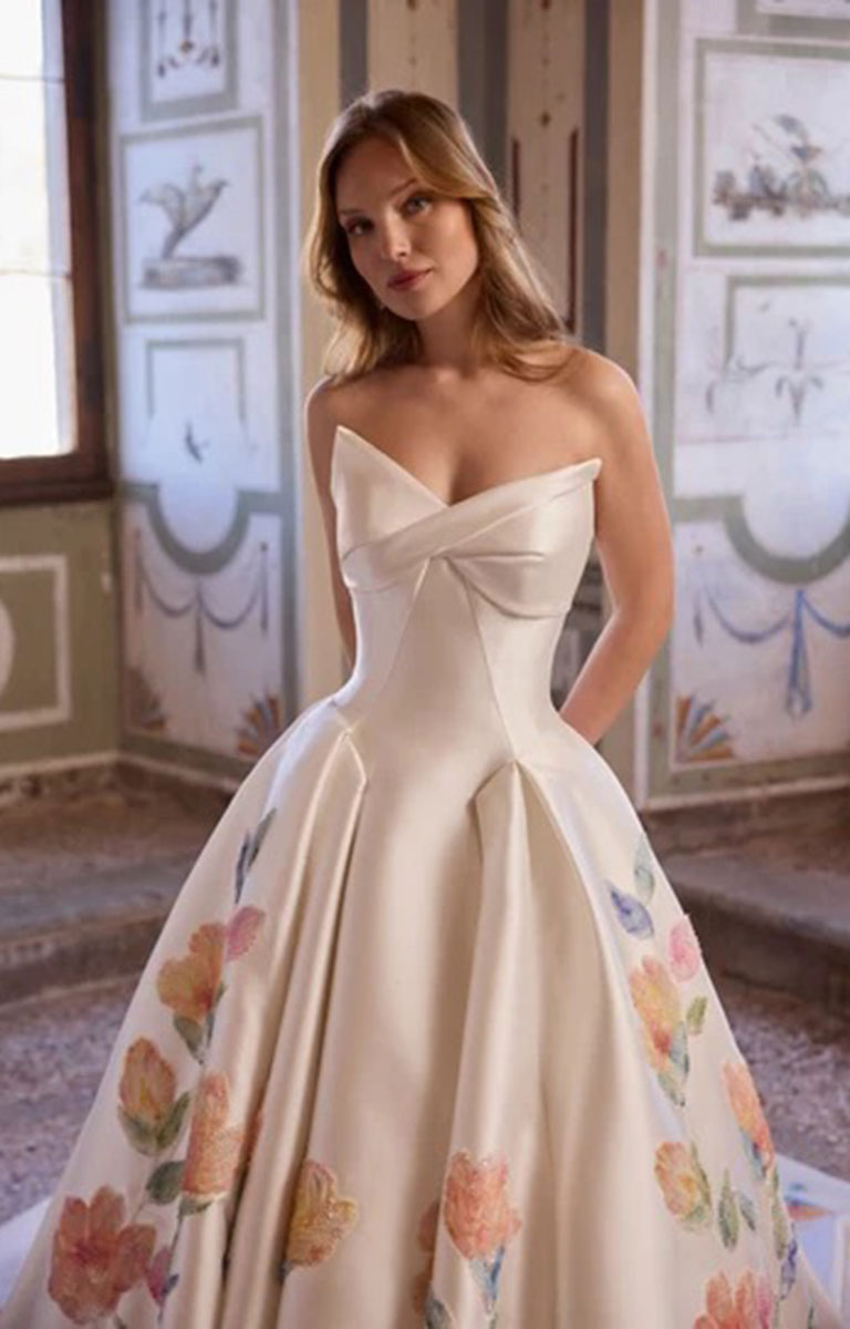 A woman models Veni Infantino Arabella 63364 - Strapless Floral Ballgown Wedding Dress with Pleated Skirt with a sweetheart neckline, a large bow at the bust, and a voluminous skirt decorated with pastel floral prints, set against an ornate interior wall.