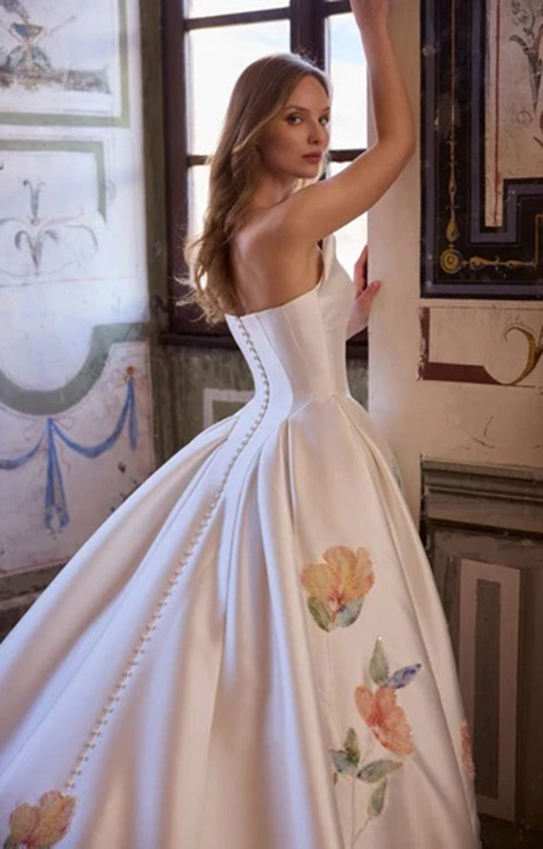A woman wearing Veni Infantino Arabella 63364 - Strapless Floral Ballgown Wedding Dress with Pleated Skirt with floral watercolor prints and a corseted bodice, posing in an ornate interior room.