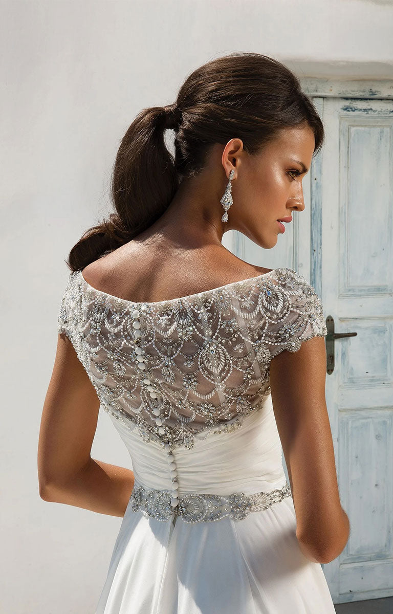 Woman wearing 8799 by Justin Alexander - Chiffon Ball Gown with Beaded Portrait Neckline with lace detailing in a softly lit room.