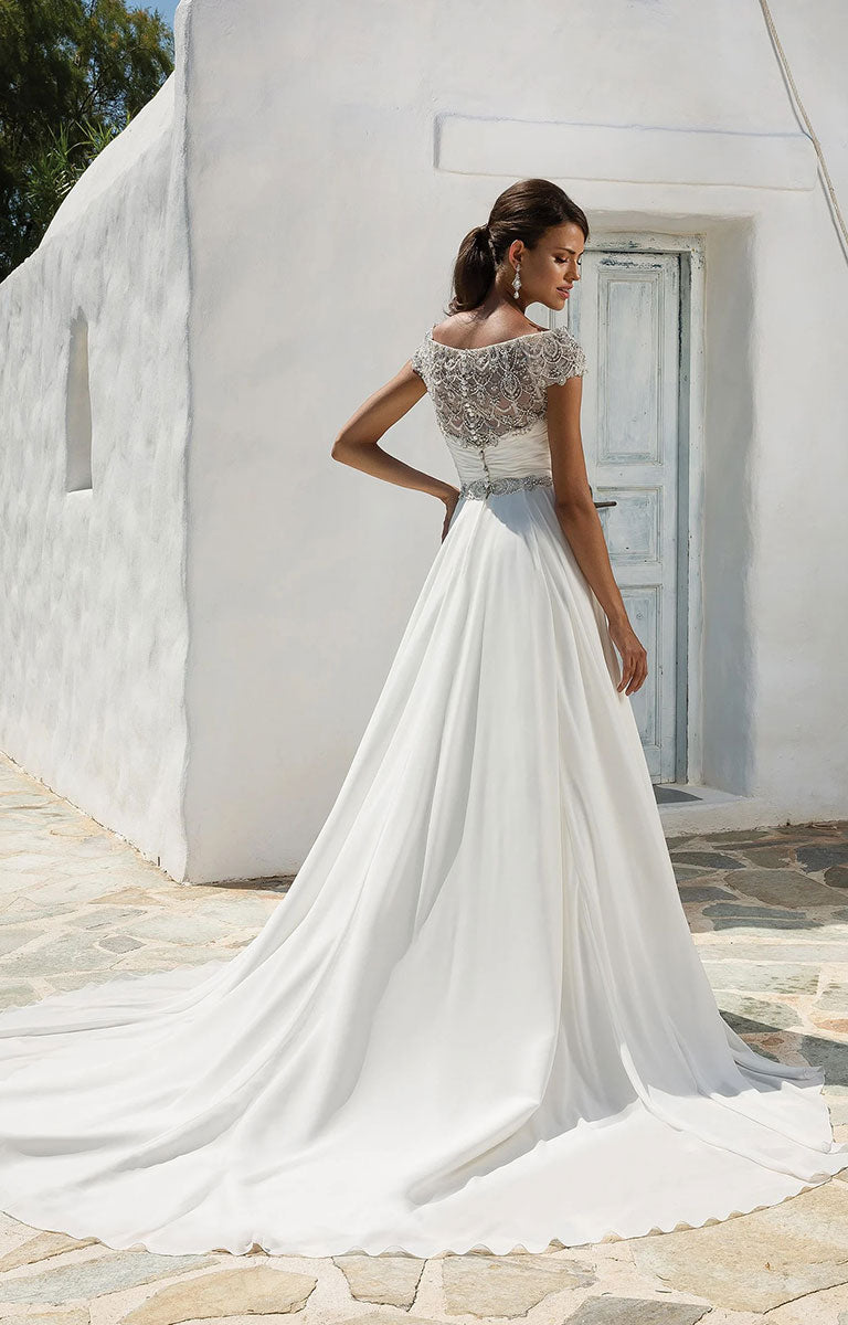 Woman in 8799 by Justin Alexander - Chiffon Ball Gown with Beaded Portrait Neckline with a textured bodice standing in front of a white building.