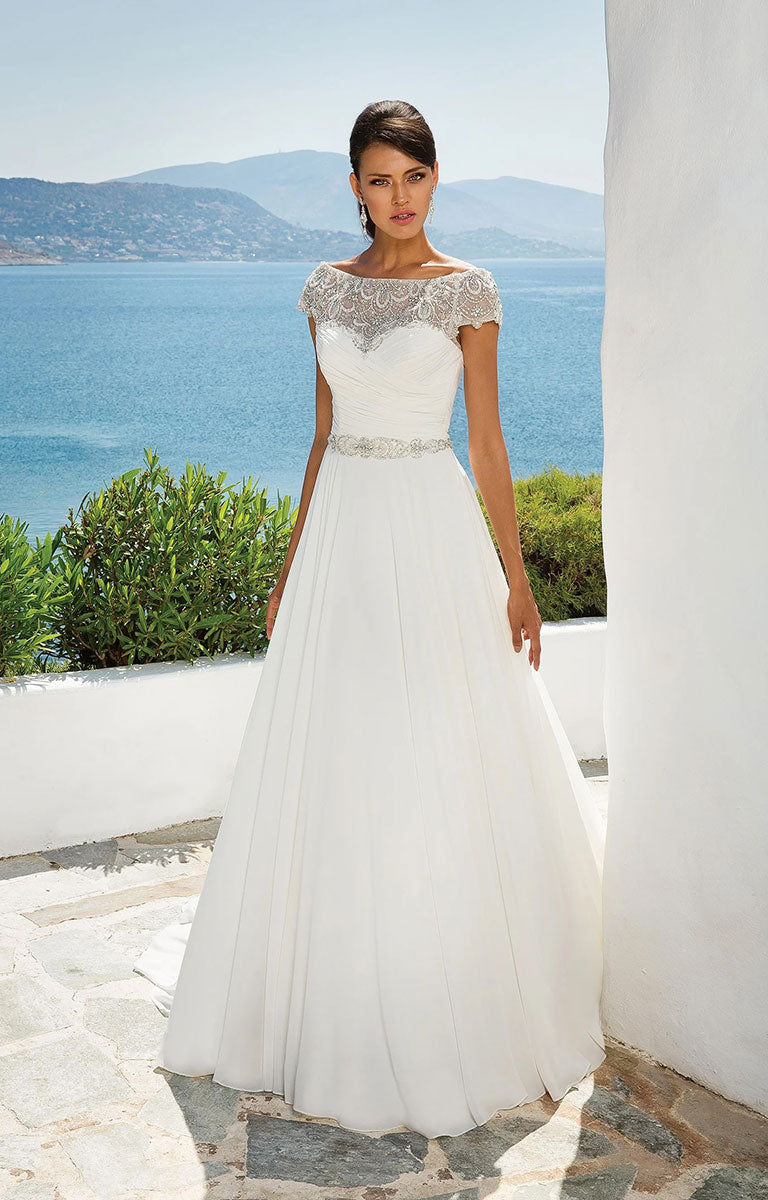 Woman in 8799 by Justin Alexander - Chiffon Ball Gown with Beaded Portrait Neckline with a scenic background of water and mountains.