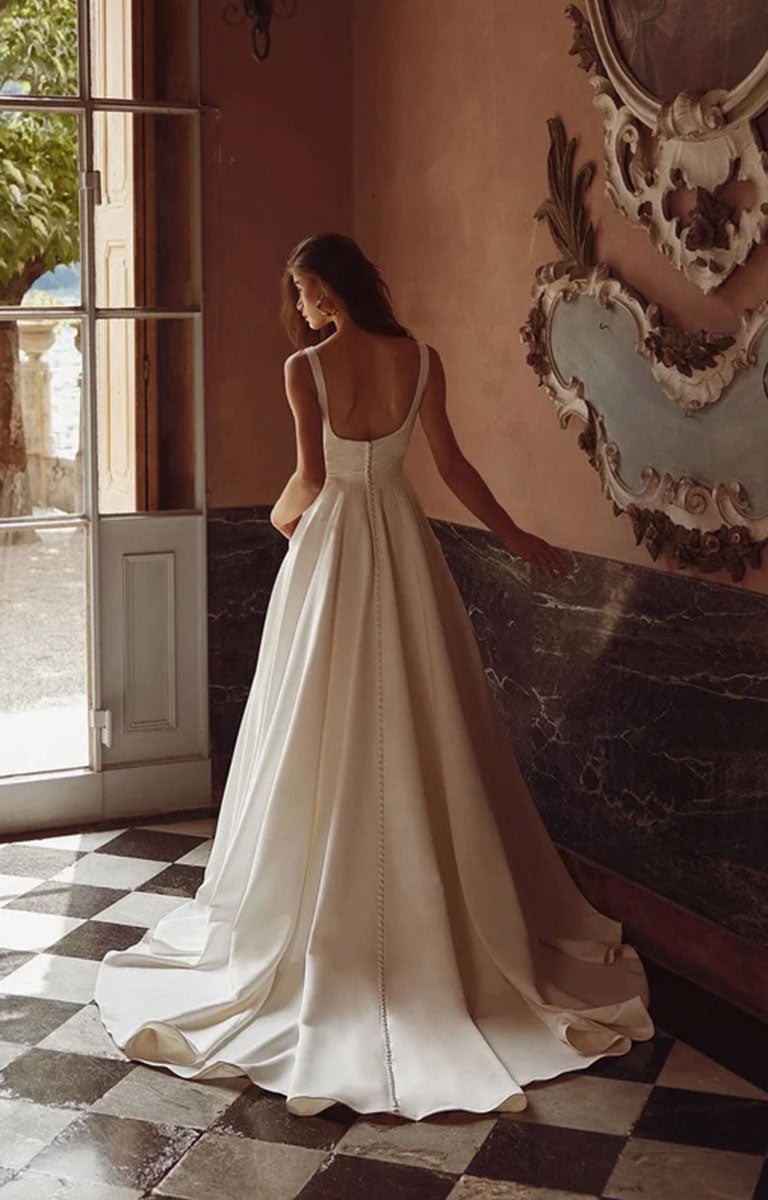 Woman in Iva by Justin Alexander - Satin Ballgown Wedding Dress with Square Neckline, Asymmetric Bodice and Pockets standing in a room with checkered floor and decorative wall.