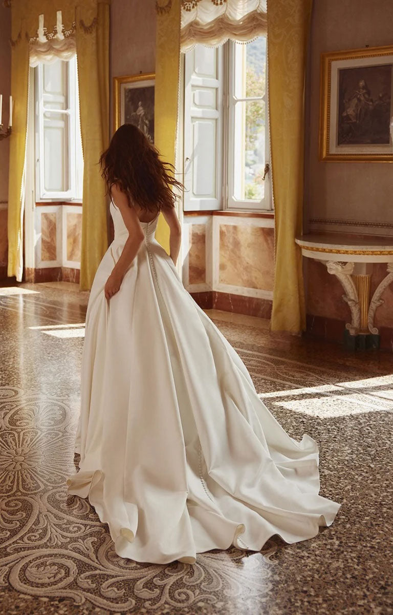 Woman in Iva by Justin Alexander - Satin Ballgown Wedding Dress with Square Neckline, Asymmetric Bodice and Pockets standing in an elegant room with yellow curtains and patterned floor.