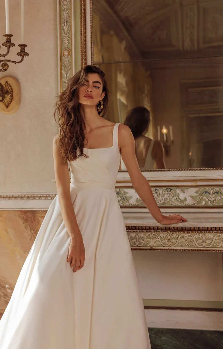 Woman in Iva by Justin Alexander - Satin Ballgown Wedding Dress with Square Neckline, Asymmetric Bodice and Pockets standing in an ornate room with a mirror.