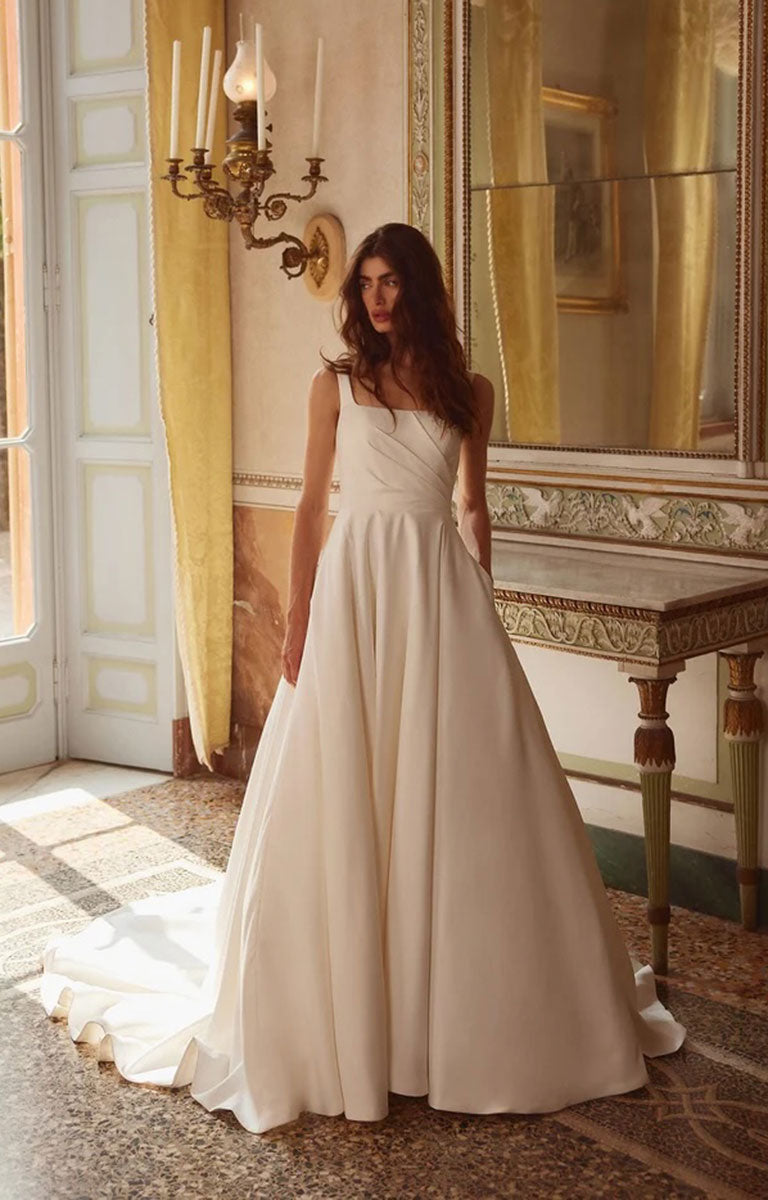 Woman in Iva by Justin Alexander - Satin Ballgown Wedding Dress with Square Neckline, Asymmetric Bodice and Pockets standing in an elegant room with a chandelier and mirror.