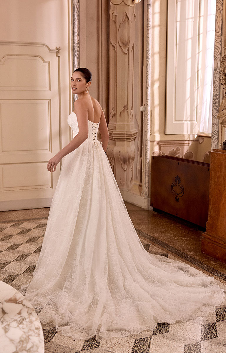 Woman in Leighton 88468 by Justin Alexander - Corded Lace A-Line Wedding Dress with Basque Waist standing in an elegant room with wooden furniture and large windows.