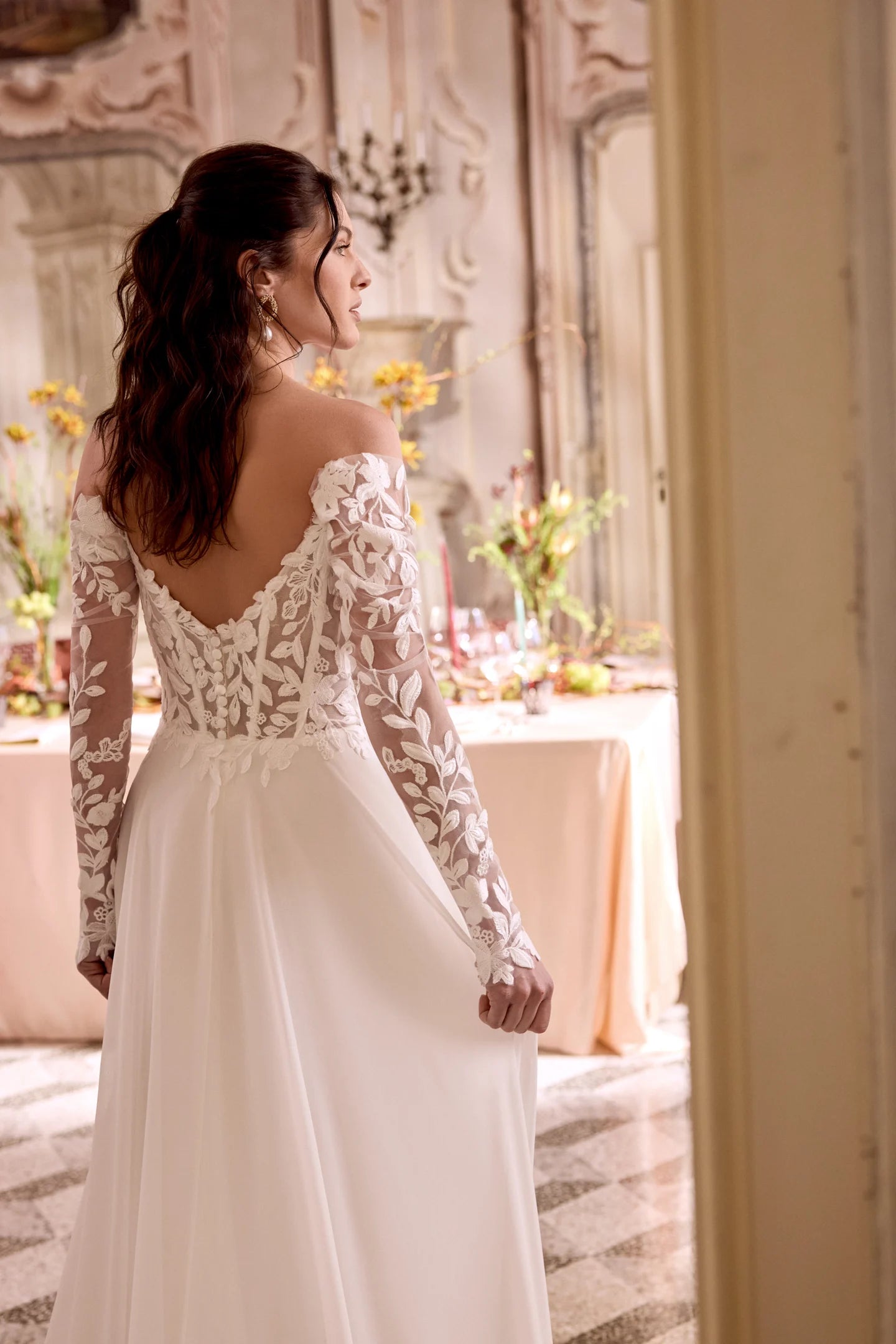Woman in a white lace wedding dress standing in an elegant room with floral arrangements.