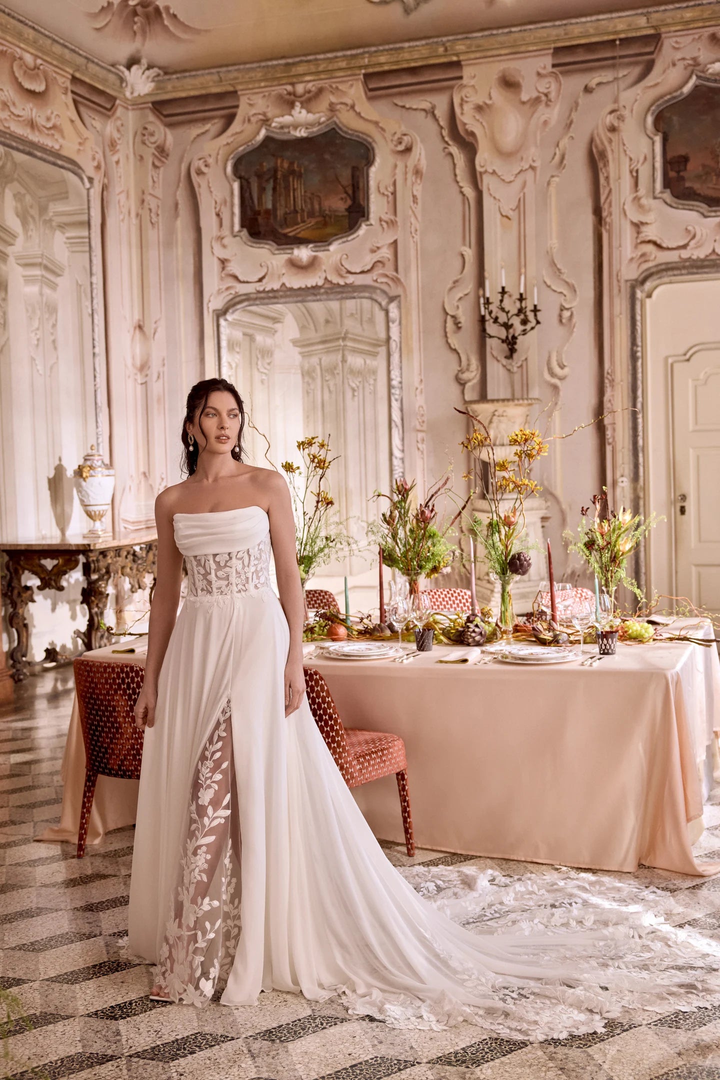 Woman in a white strapless gown standing in an ornate room with a table set for a meal.