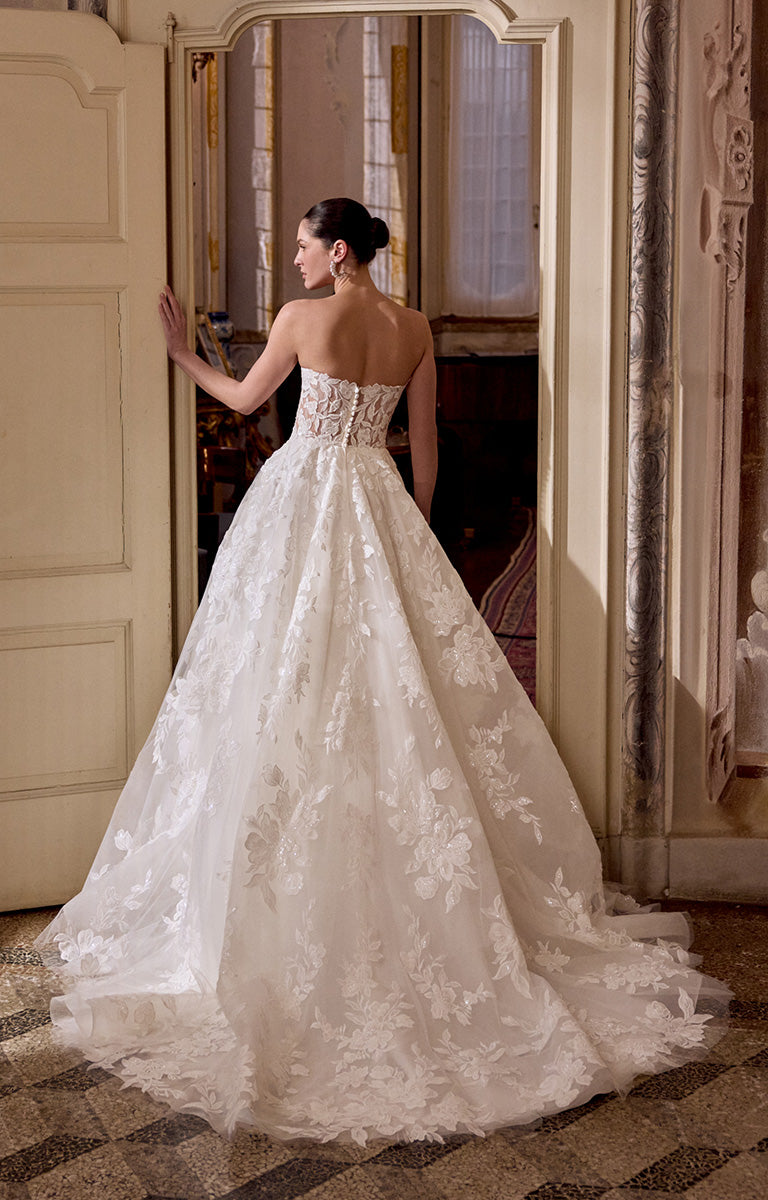 Woman in a white lace wedding dress standing in a room with ornate decor.