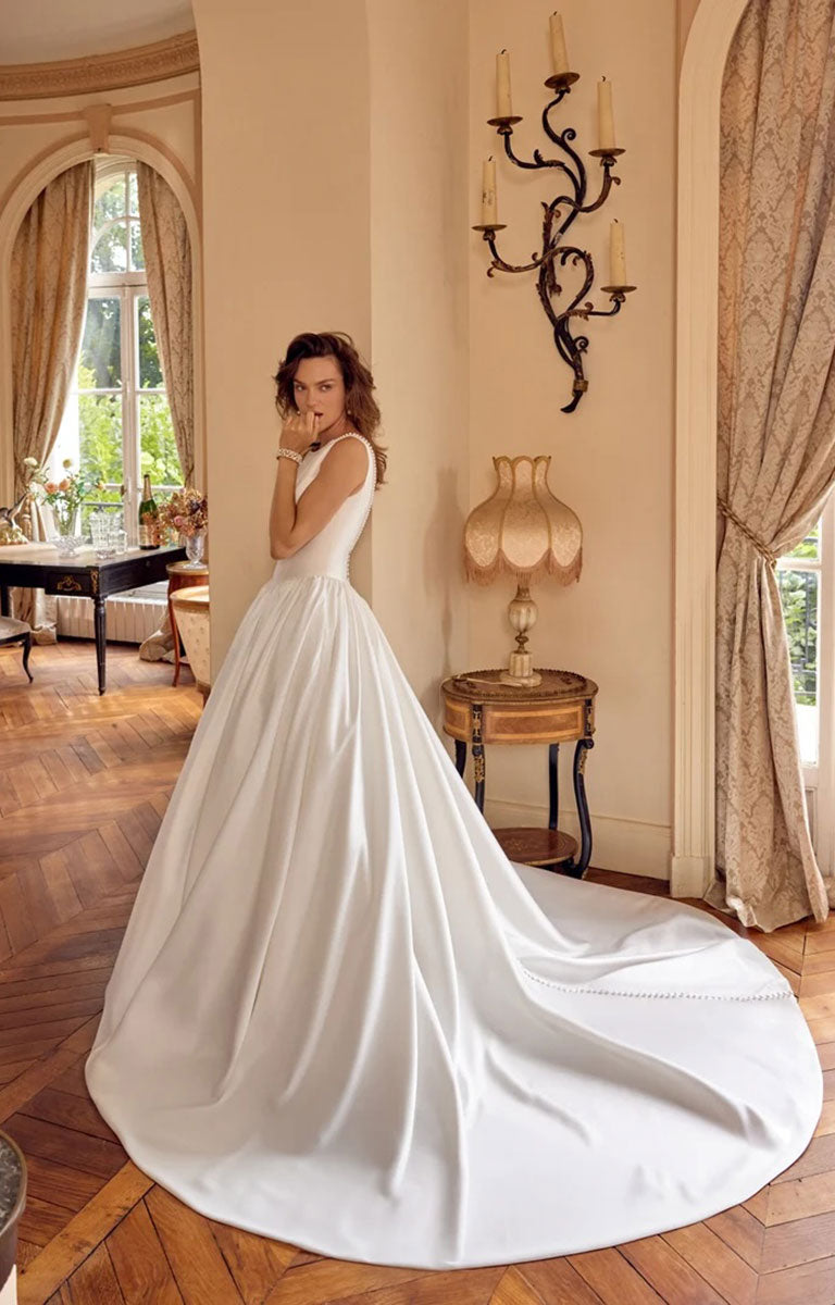 A woman wearing Mckenna by Justin Alexander - Mikado Ball Gown with Pearl-Trimmed Sabrina Neckline and Low V-Back with a long train, standing in an elegant room with wood floors, curtains, and a wall sconce.