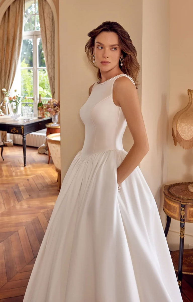 A woman modeling Mckenna by Justin Alexander - Mikado Ball Gown with Pearl-Trimmed Sabrina Neckline and Low V-Back with a fitted bodice, high neckline, and full skirt in an elegant indoor room with wood floors and warm decor.