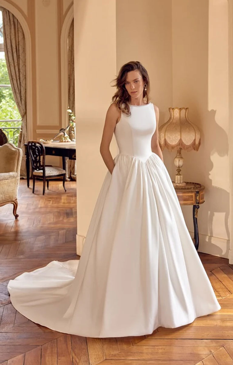 A woman modeling Mckenna by Justin Alexander - Mikado Ball Gown with Pearl-Trimmed Sabrina Neckline and Low V-Back with a fitted bodice and full skirt in an elegant indoor room with wood floors and warm beige walls.