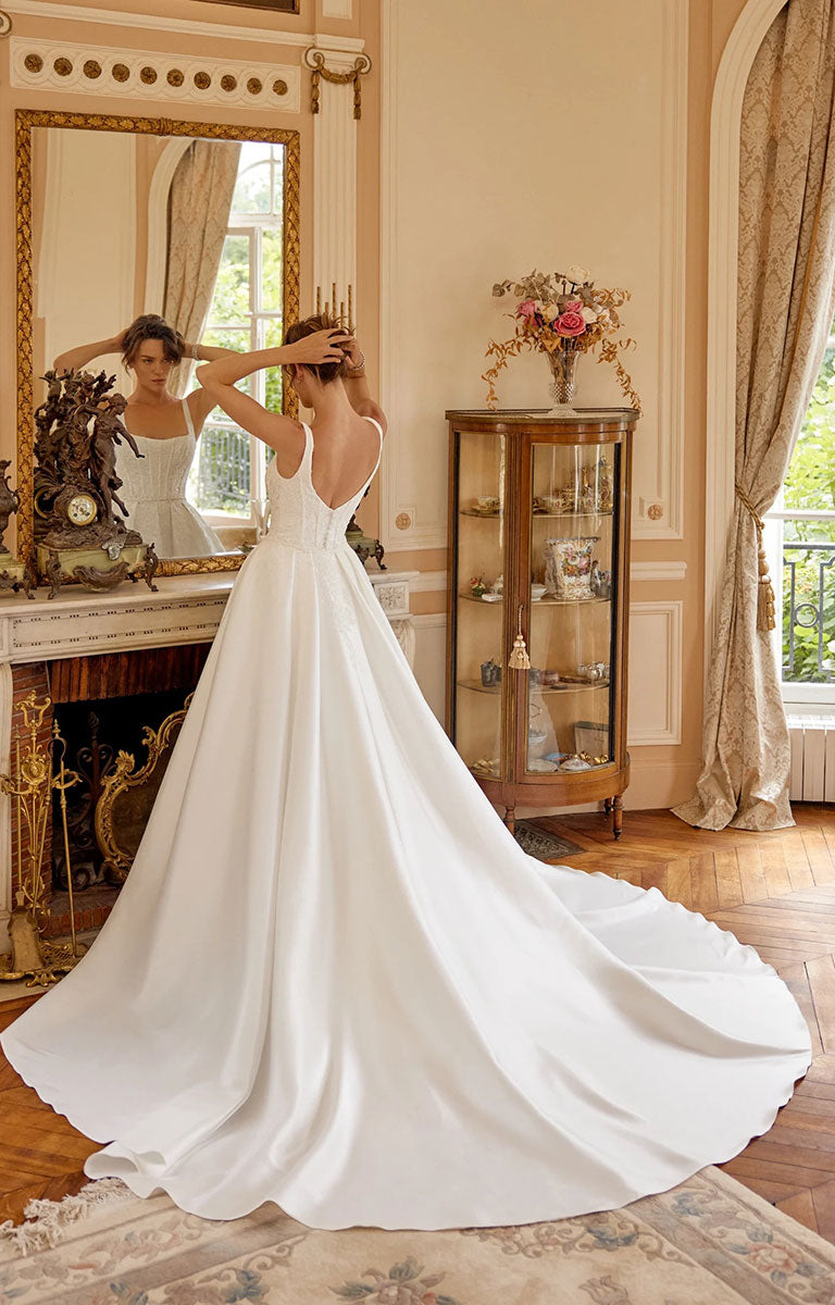 A woman wearing Marley 88519 by Justin Alexander - Stretch Mikado Fit-and-Flare Wedding Dress with Lace Appliqués and Optional Overskirt with a long train stands in a grand room, facing a large mirror that reflects her from the back. The room has warm beige walls, a fireplace, a wooden display cabinet, and curtains by a window.