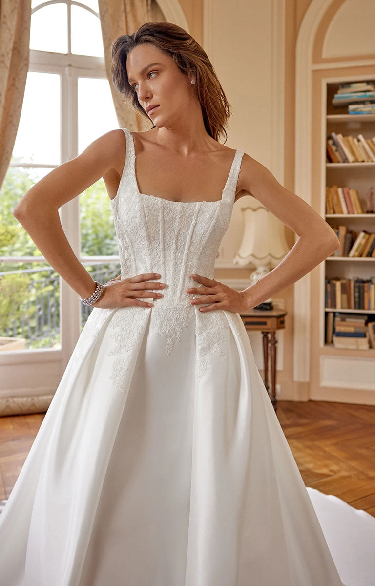 A woman modeling Marley 88519 by Justin Alexander - Stretch Mikado Fit-and-Flare Wedding Dress with Lace Appliqués and Optional Overskirt with a structured lace bodice and full satin skirt, standing in a warm, elegant room with bookshelves and a window.