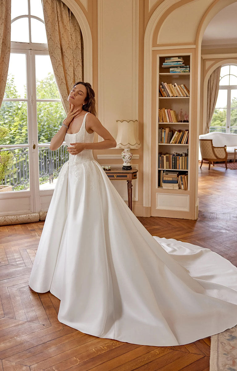 A woman wearing Marley 88519 by Justin Alexander - Stretch Mikado Fit-and-Flare Wedding Dress with Lace Appliqués and Optional Overskirt, standing in a classic French-style room with wooden floors, bookshelves, and tall windows.