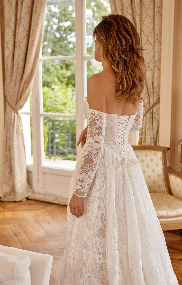 A woman wearing Magnolia 88517 by Justin Alexander - Corded Alençon Lace A-Line Wedding Dress with Sweetheart Neckline stands with her back to the camera in a sunlit room with large windows, curtains, and a wooden floor.