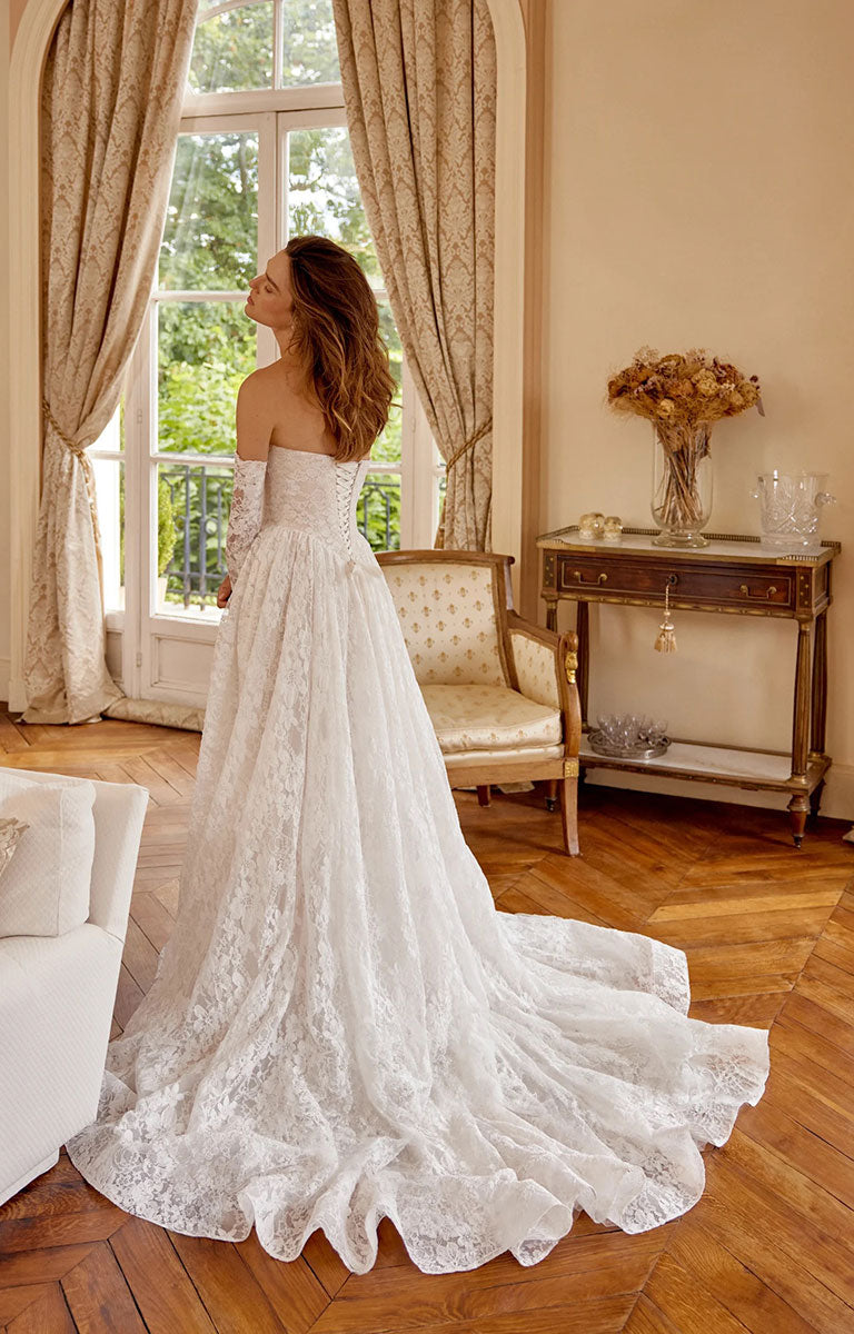 A woman wearing Magnolia 88517 by Justin Alexander - Corded Alençon Lace A-Line Wedding Dress with Sweetheart Neckline with a long train stands in a sunlit room with wood floors, beige curtains, and a small table in the background.