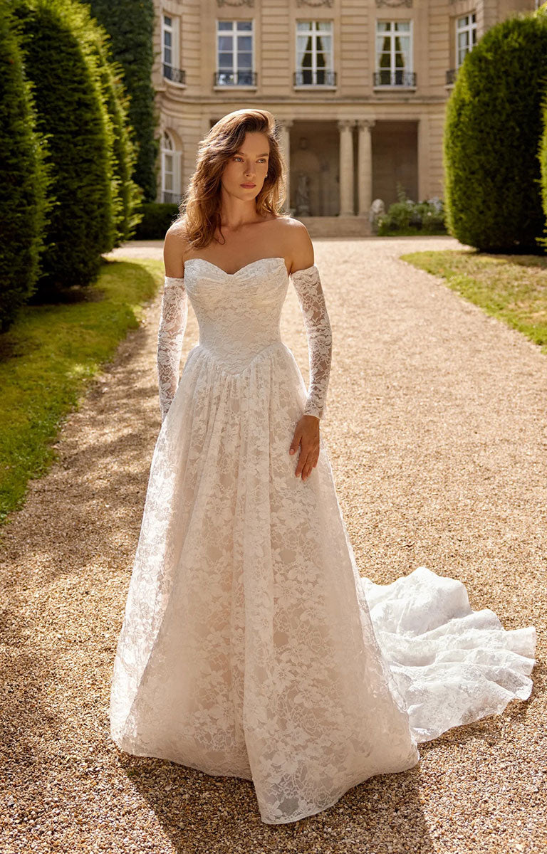 A woman wearing Magnolia 88517 by Justin Alexander - Corded Alençon Lace A-Line Wedding Dress with Sweetheart Neckline with long sheer sleeves and a full skirt stands on a gravel path in front of a grand chateau.