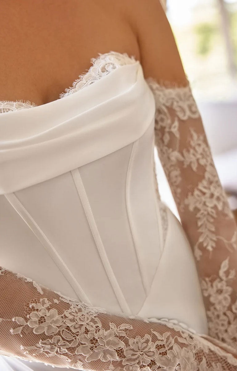 Close-up of Mimi by Justin Alexander - Charmeuse Fit-and-Flare Wedding Dress with Exposed Boning and Basque Waist with long sheer lace sleeves, showing the satin-like bodice and floral lace details against a softly blurred light background.