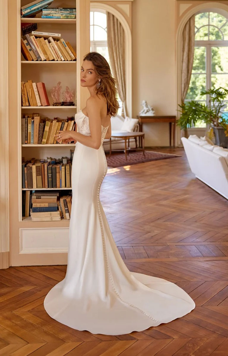 A woman wearing Mimi by Justin Alexander - Charmeuse Fit-and-Flare Wedding Dress with Exposed Boning and Basque Waist with a fitted bodice, sheer upper section, and a long train stands in a bright room with wood floors and bookshelves.