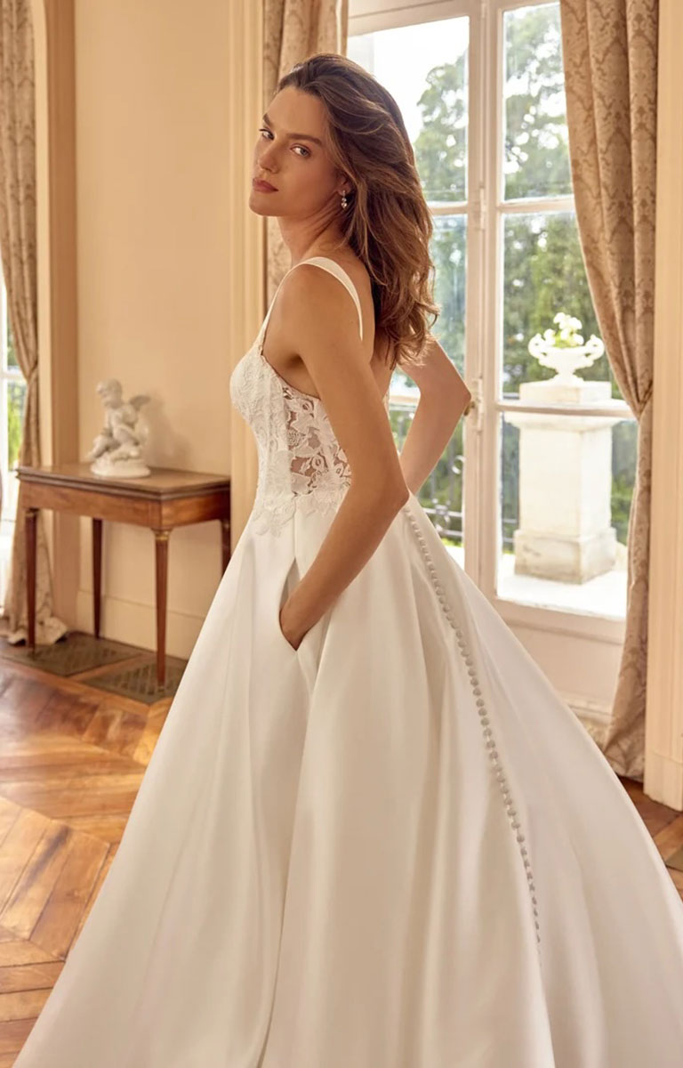 A woman modeling Matilda by Justin Alexander - Mikado A-Line Wedding Dress with Sweetheart Neckline and Matte Lace Appliqués in a bright, elegant room with large windows, wood floors, and beige curtains. The dress features a lace bodice, thin straps, and a full skirt with a decorative beaded side detail.