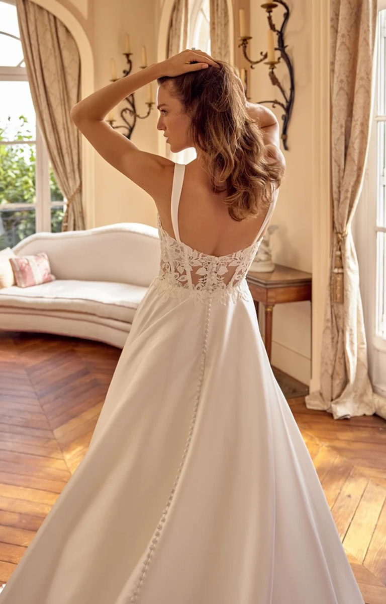A woman wearing Matilda by Justin Alexander - Mikado A-Line Wedding Dress with Sweetheart Neckline and Matte Lace Appliqués with a lace-embellished open back, photographed from behind in a bright, elegant room with wood floors and cream curtains.