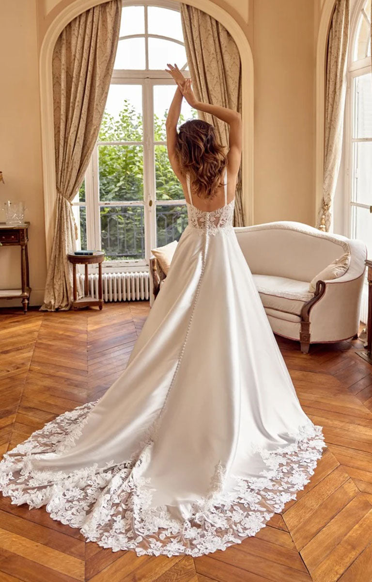 A woman wearing Matilda by Justin Alexander - Mikado A-Line Wedding Dress with Sweetheart Neckline and Matte Lace Appliqués with a lace-trimmed train, standing in a sunlit room with wood floors, arched windows, and a cream sofa.