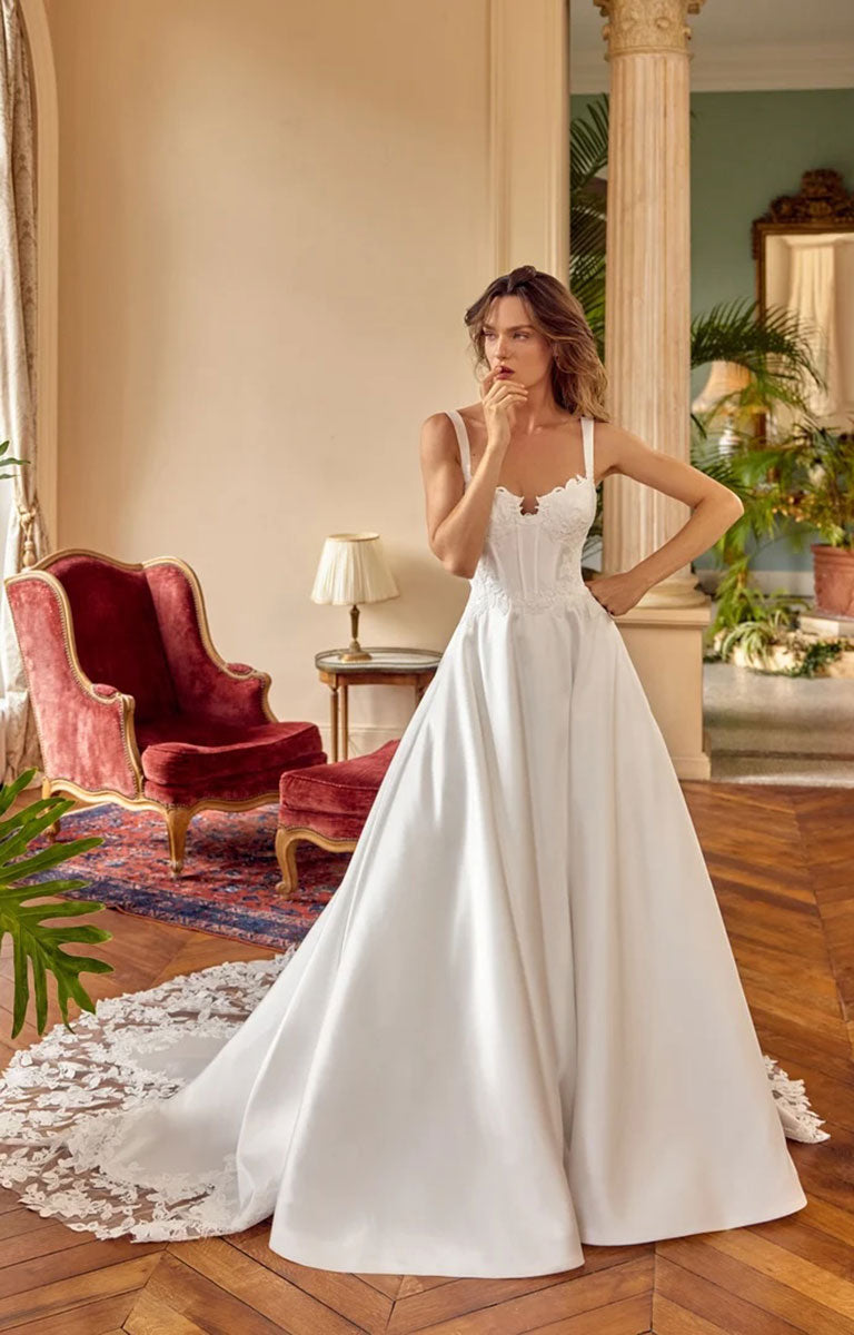 A woman modeling Matilda by Justin Alexander - Mikado A-Line Wedding Dress with Sweetheart Neckline and Matte Lace Appliqués with a lace-trimmed sweetheart bodice and a long train, standing in an elegant room with wood floors, a red velvet chair, and a lamp.