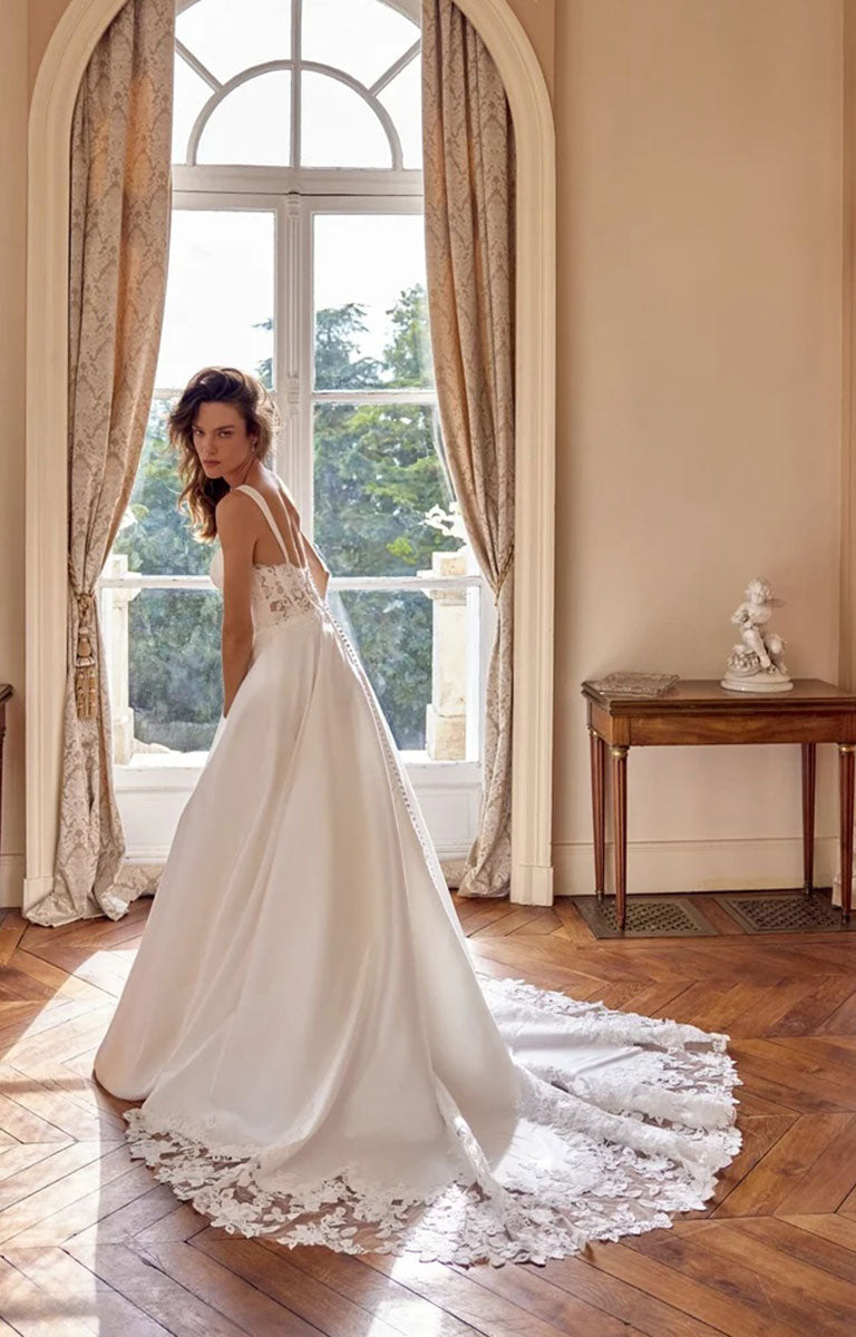 A woman wearing Matilda by Justin Alexander - Mikado A-Line Wedding Dress with Sweetheart Neckline and Matte Lace Appliqués stands in a sunlit room with tall windows, patterned curtains, and a wood floor, looking back over her shoulder as the dress train spreads across the floor.