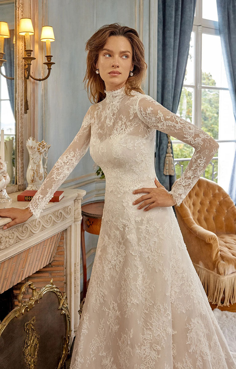 A woman wearing Merida 88525 by Justin Alexander - Alençon Lace A-Line Wedding Dress with Eyelash Trim with a high mock neck, standing in an elegant room with a fireplace, mirror, and window.