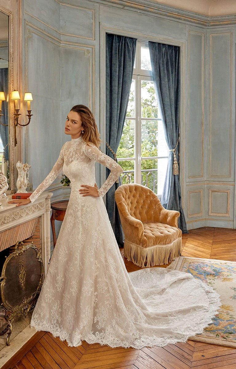 A woman wearing Merida 88525 by Justin Alexander - Alençon Lace A-Line Wedding Dress with Eyelash Trim with a high mock neck and a long train, standing in an elegant room with blue paneled walls, a window, and a gold armchair.
