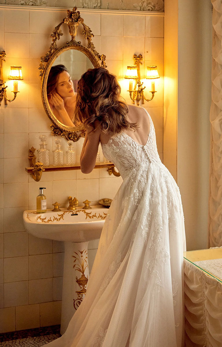 A woman in Melina 88529 by Justin Alexander - Sparkle Tulle A-Line Wedding Dress with Plunging V-Neck stands in a warmly lit bathroom, looking into an ornate gold mirror above a pedestal sink.