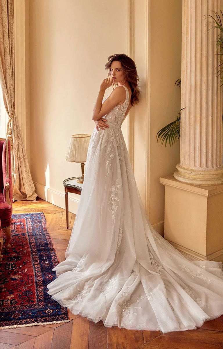 A woman wearing Melina 88529 by Justin Alexander - Sparkle Tulle A-Line Wedding Dress with Plunging V-Neck with a long train, standing in a warm, elegant room with cream walls, a red patterned rug, and a column.