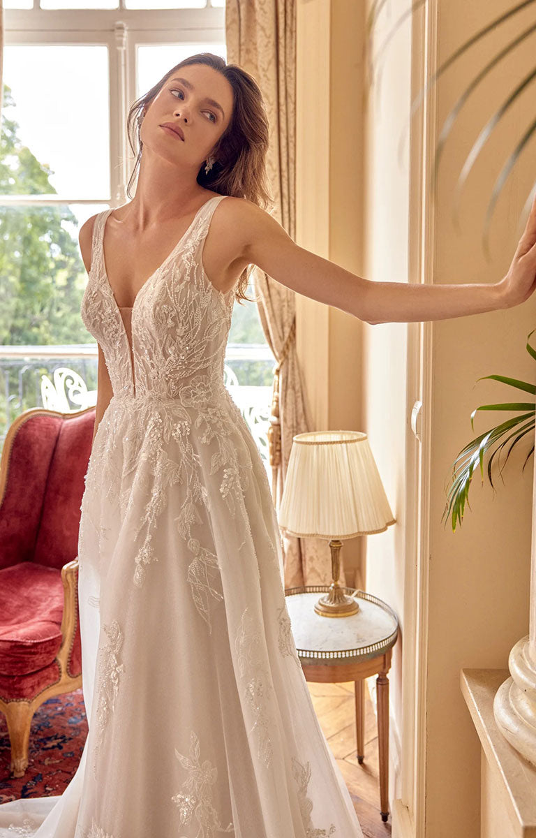 A woman wearing Melina 88529 by Justin Alexander - Sparkle Tulle A-Line Wedding Dress with Plunging V-Neck with a deep V-neckline and full skirt, posed indoors near a window with a red armchair, lamp, and plant in the background.