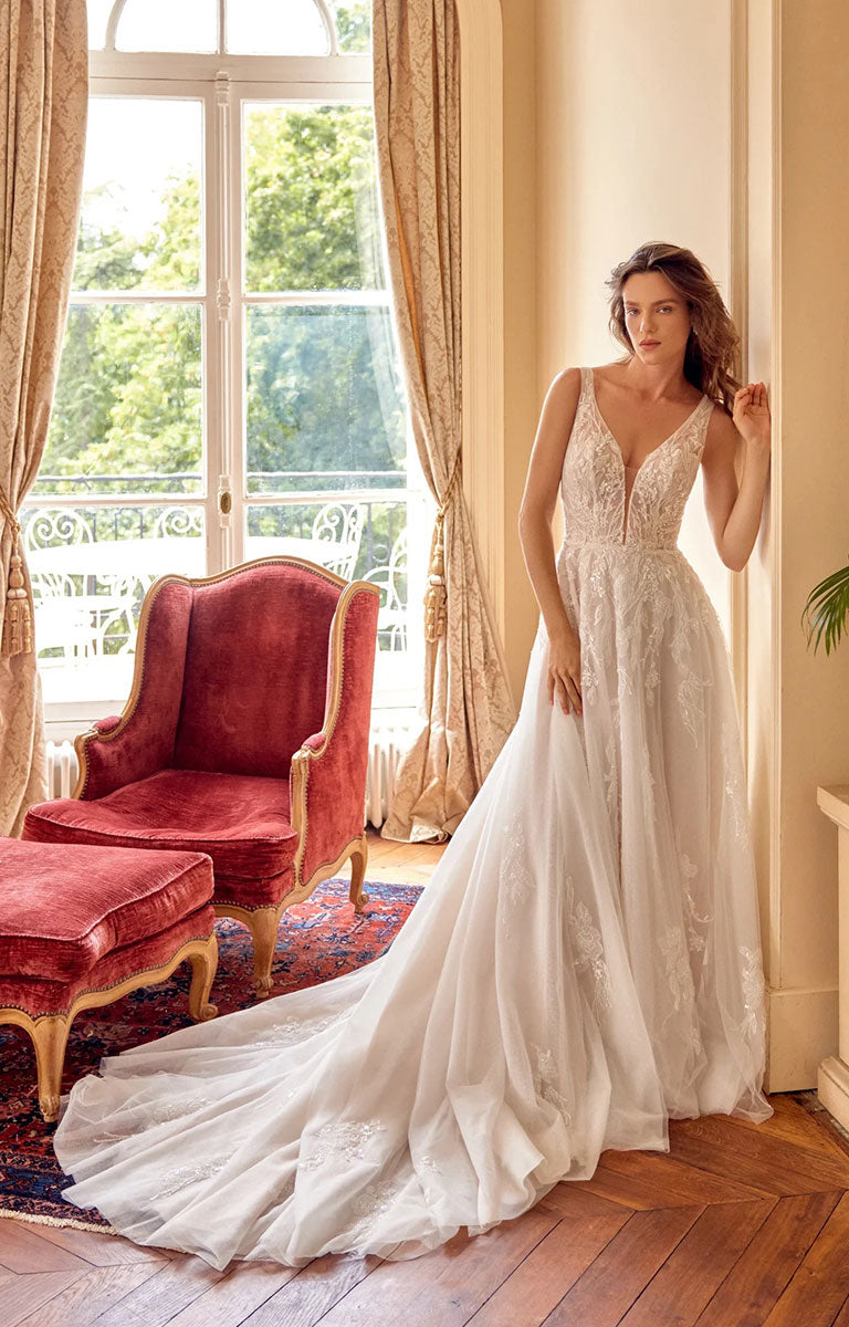 A woman wearing Melina 88529 by Justin Alexander - Sparkle Tulle A-Line Wedding Dress with Plunging V-Neck with a long train stands in a sunlit room beside a red velvet armchair and window.