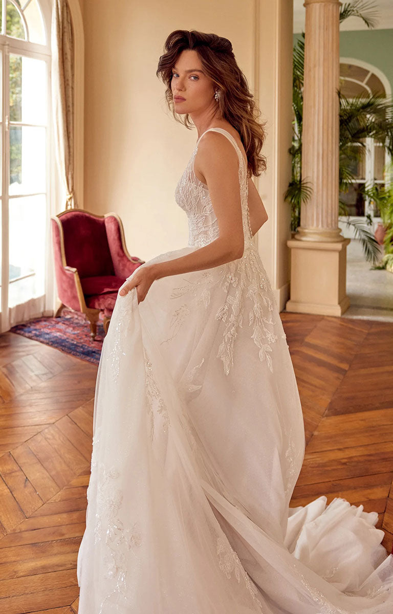 A woman wearing Melina 88529 by Justin Alexander - Sparkle Tulle A-Line Wedding Dress with Plunging V-Neck with lace and tulle detailing, standing in a grand interior room with wood floors, columns, and a red armchair.
